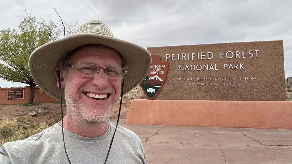 Shea wearing a wide-brim hat smiles in front of the Petrified Forest National Park entrance sign under a cloudy sky.