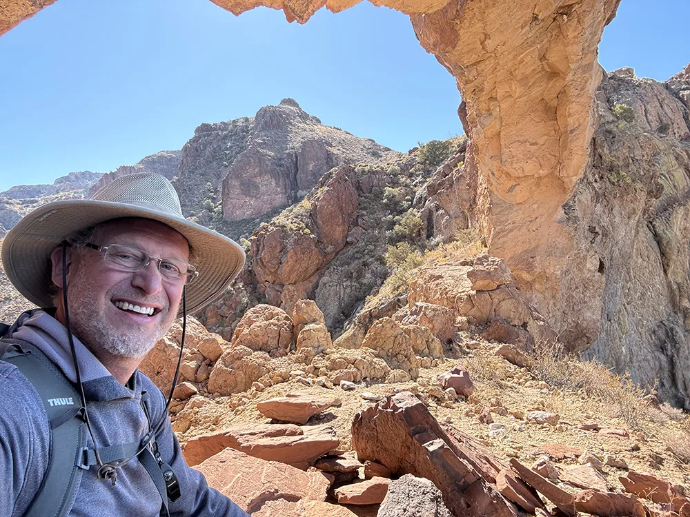Shea Oliver wearing a hat and glasses smiles at the camera while hiking in a rocky desert landscape with a natural rock arch overhead, contemplating ideas for Affiliate Marketing and Building a Profitable Online Business.