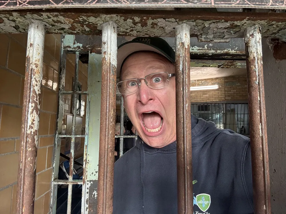 Shea wearing glasses and a cap yells with a surprised expression while standing behind rusty metal jail bars indoors.