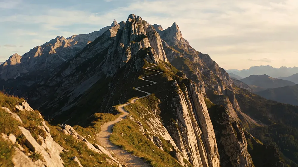 A narrow dirt path zigzags up a steep, rocky mountain ridge under a partly cloudy sky, with distant peaks visible in the background—much like the journey to start a profitable blog and begin making money online.